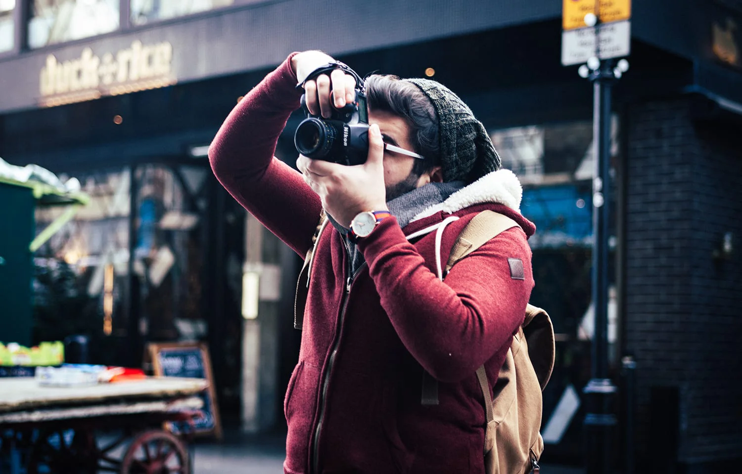 A person captures moments with a camera in an urban setting, dressed in a cozy red jacket and beanie.