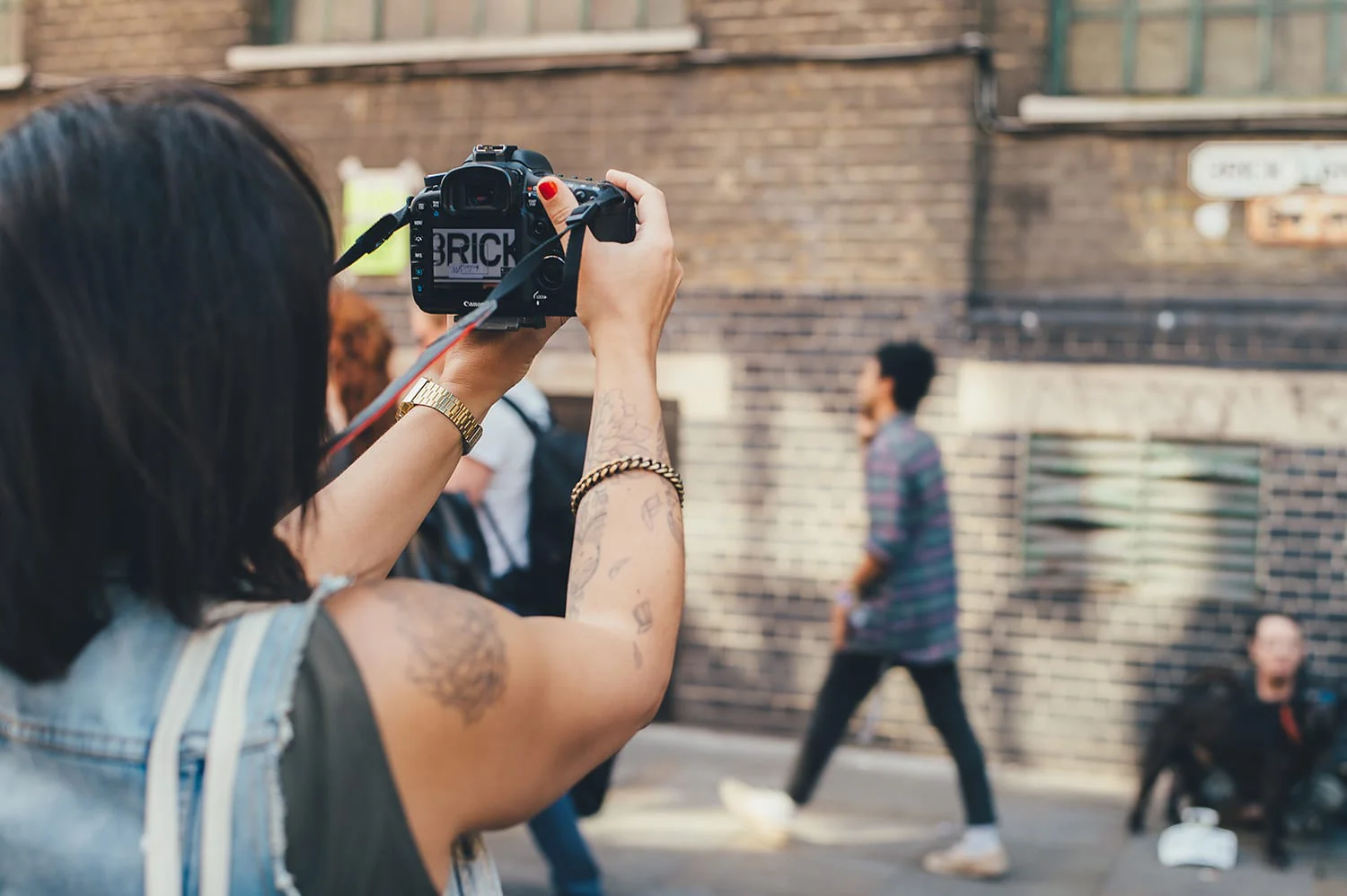 A photographer uses a digital SLR camera to capture an urban scene. People are seen moving in the background, and there is a brick environment.