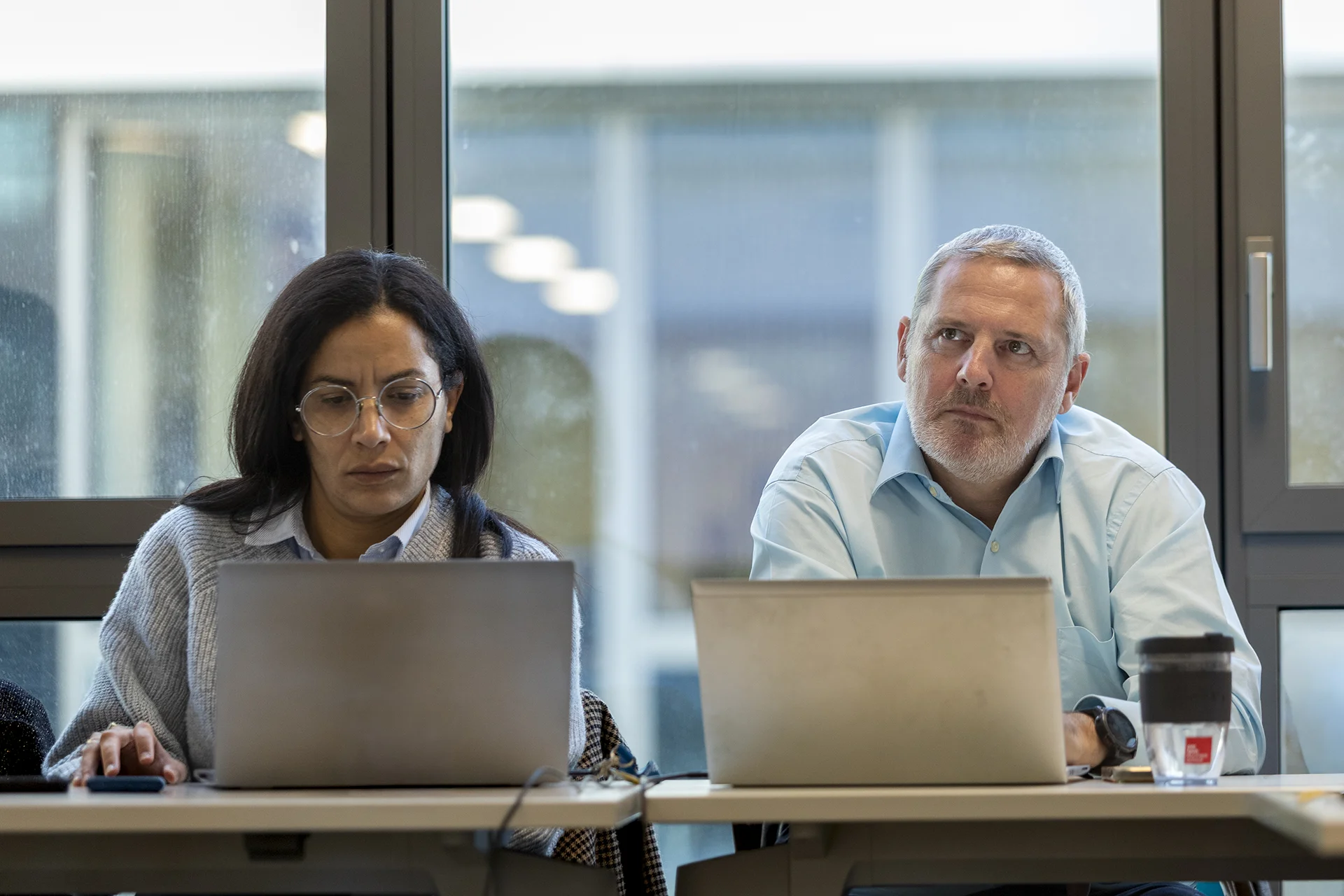 Deux personnes assises à une table de réunion, travaillant sur leurs ordinateurs portables. Un café et un verre d'eau sont présents sur la table, avec une grande fenêtre en arrière-plan laissant passer la lumière.