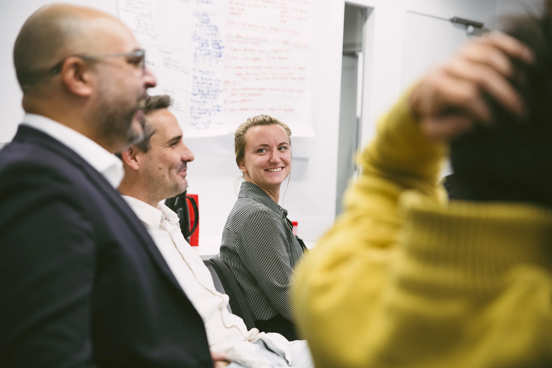 Dans cette image, on voit un groupe de personnes assises dans une salle, vraisemblablement lors d'une réunion ou d'une séance de brainstorming. On aperçoit des notes accrochées au mur en arrière-plan, ce qui indique qu'ils pourraient discuter de divers sujets. Les participants semblent attentifs et engagés dans la conversation. Les vêtements des personnes montrent une ambiance professionnelle, et l'environnement est moderne et minimaliste.