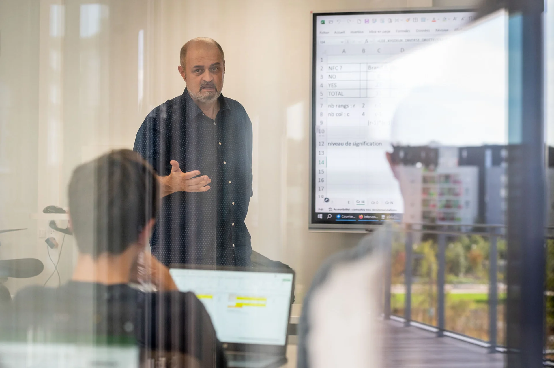 Un enseignant se tenant debout devant un tableau numérique, expliquant un sujet à des étudiants assis, chacun devant son ordinateur portable. La scène se déroule à emlyon, avec une ambiance d'apprentissage dynamique et interactive. Le tableau présente des données et des informations, suggérant un cours axé sur l'analyse ou la gestion des données.
