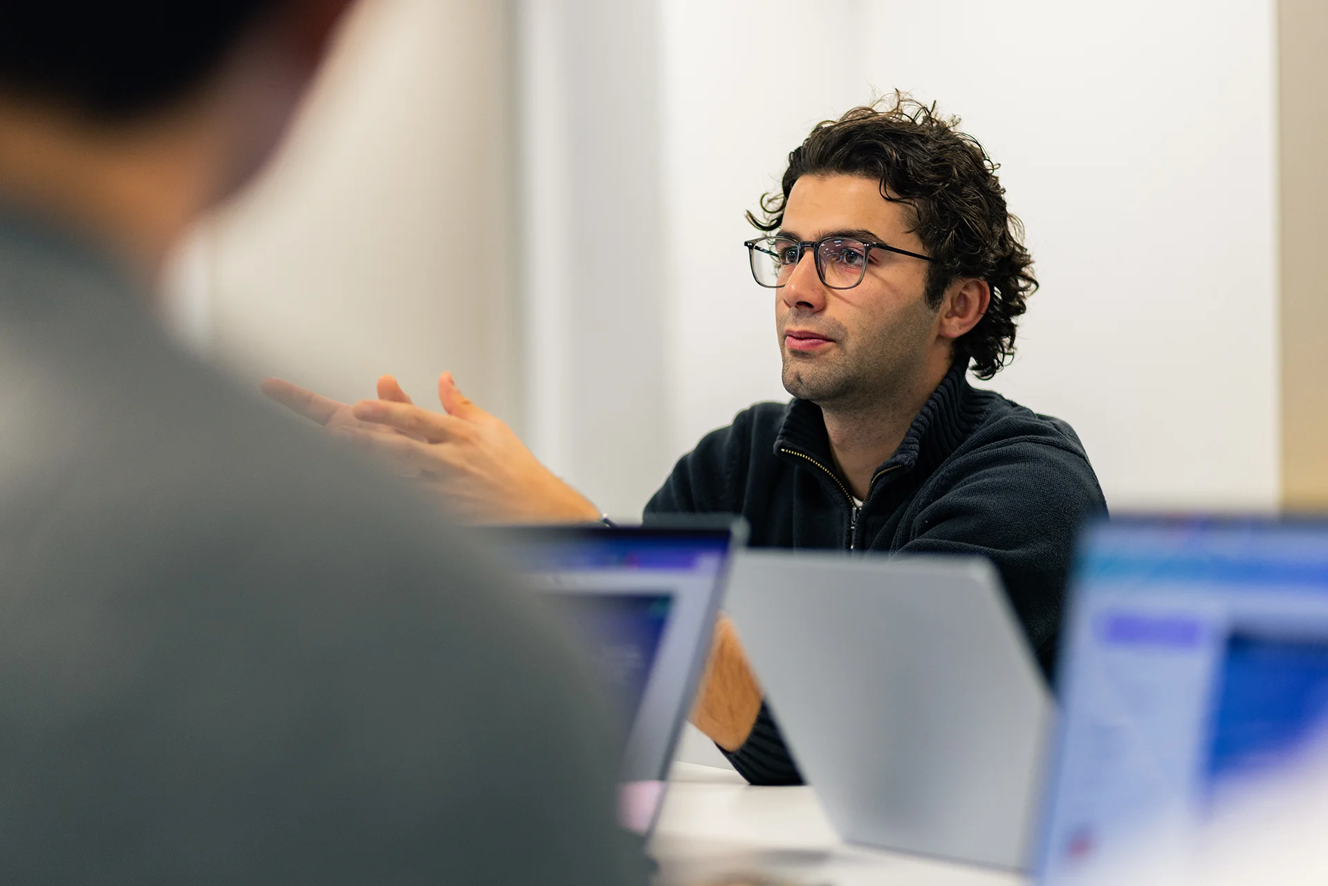 Discussion en réunion avec un participant gesticulant, portant un pull noir et des lunettes, devant des ordinateurs portables, dans un environnement de travail moderne.
