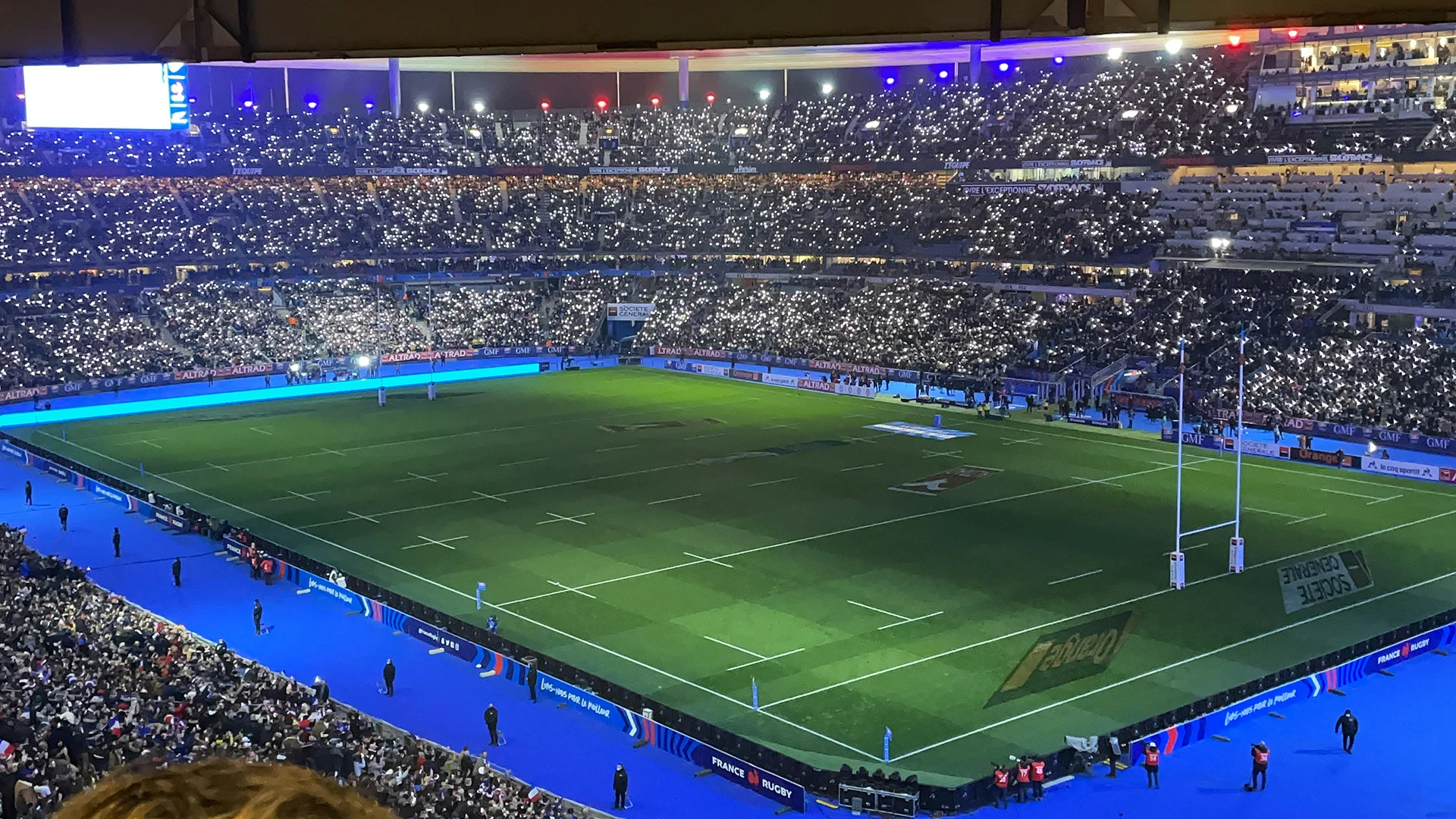 Vue du terrain de rugby avec des lumières scintillantes dans un stade rempli de supporters.