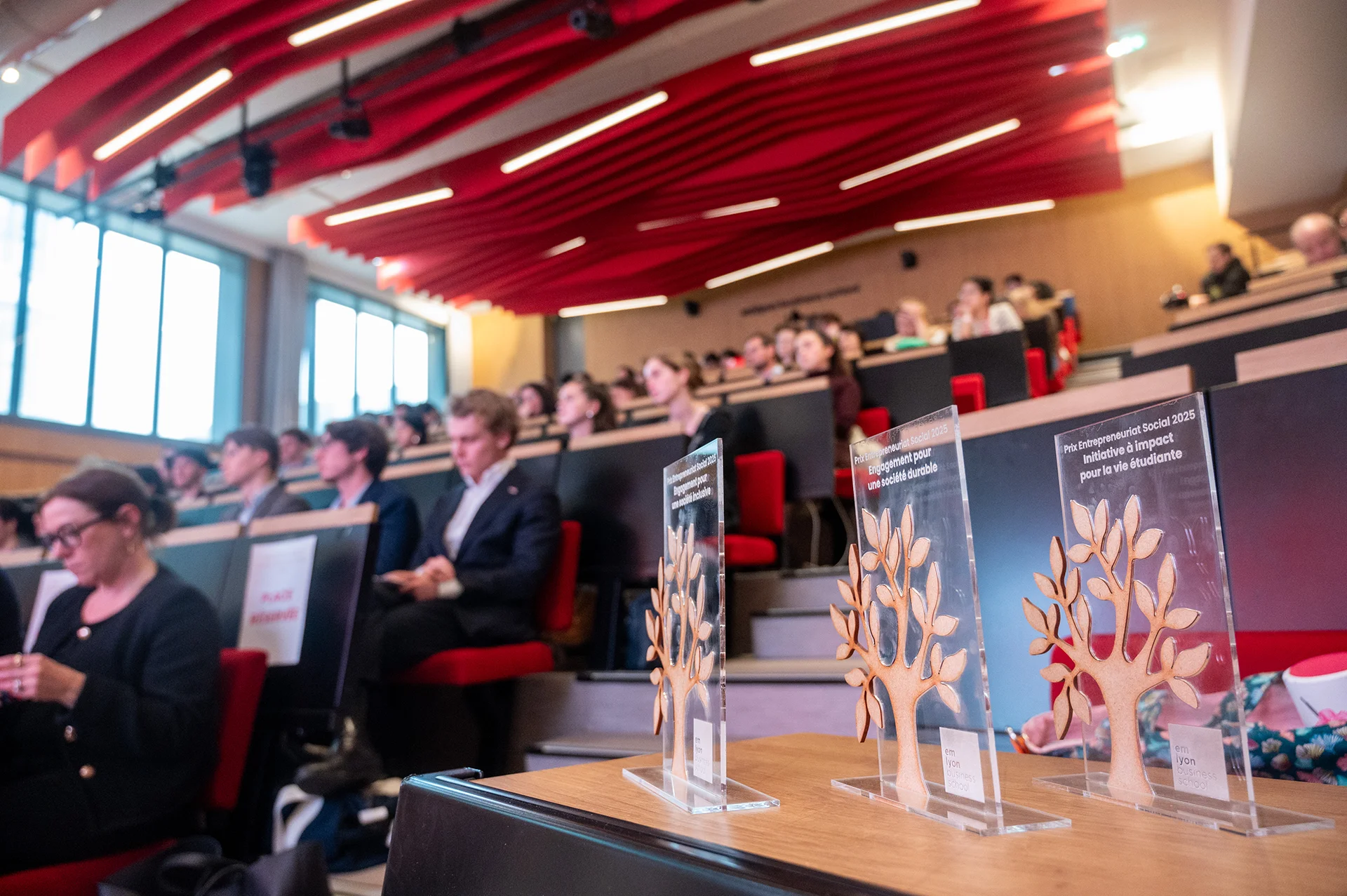 Awards displayed on a table during a ceremony in a lecture hall filled with attendees.