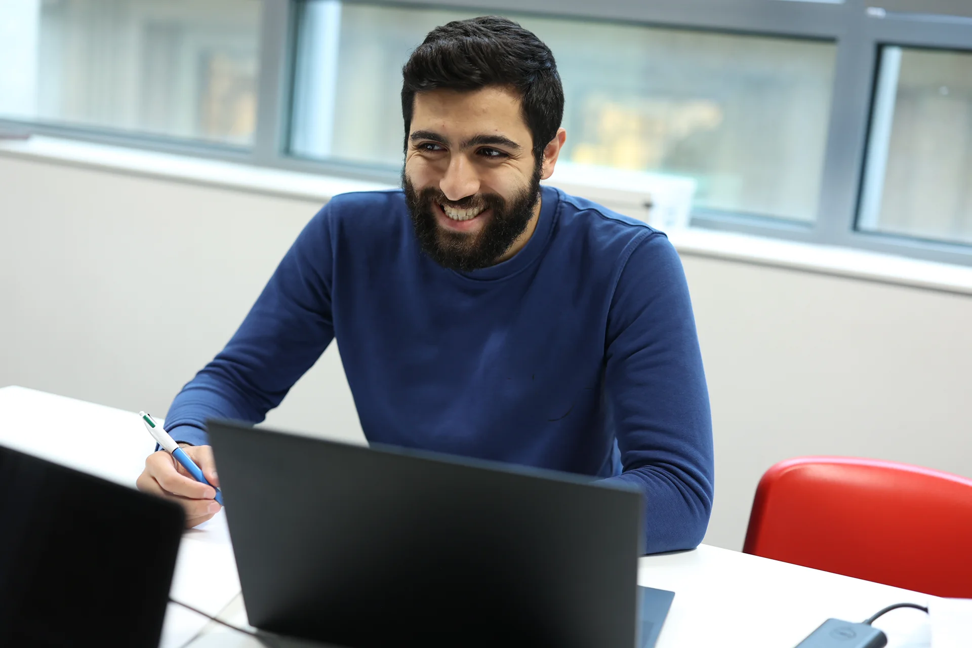 Un homme assis à une table, portant un pull bleu. Il tient un stylo et semble écrire. Sur la table, il y a un ordinateur portable noir et un autre appareil. Une fenêtre se trouve en arrière-plan.