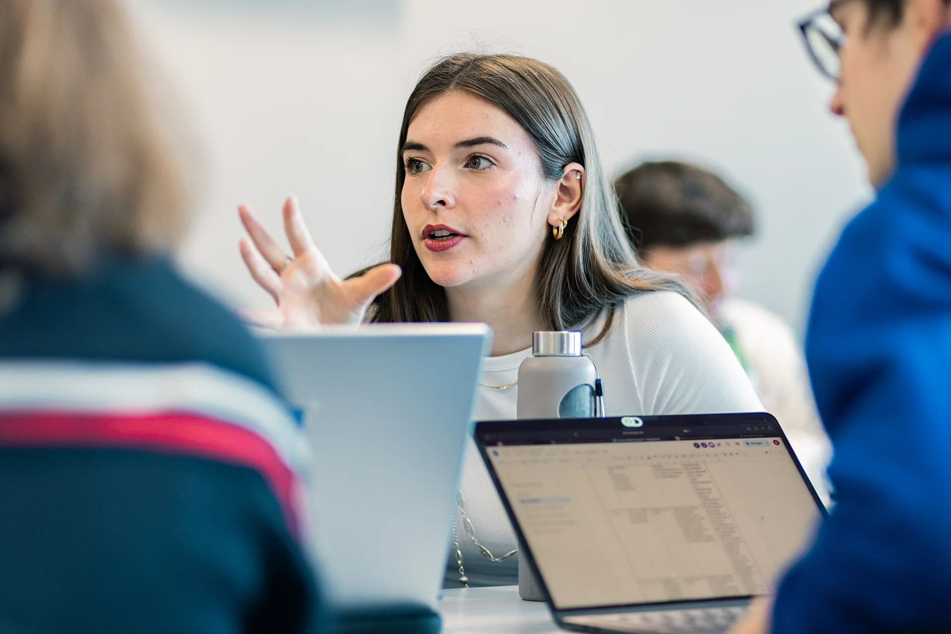 Un groupe d'étudiants en train de discuter dans un environnement d'apprentissage dynamique, avec plusieurs ordinateurs portables ouverts sur la table. L'un des étudiants utilise des gestes pour exprimer ses idées, tandis que les autres écoutent attentivement. L'atmosphère semble collaborative et engagée.