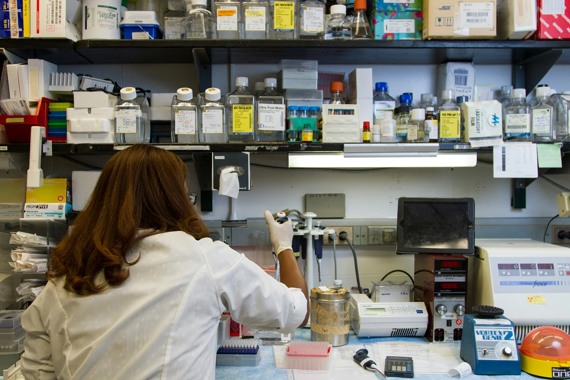 Research worker in a fully equipped laboratory handling scientific instruments on a bench filled with equipment, reagents and lab materials.