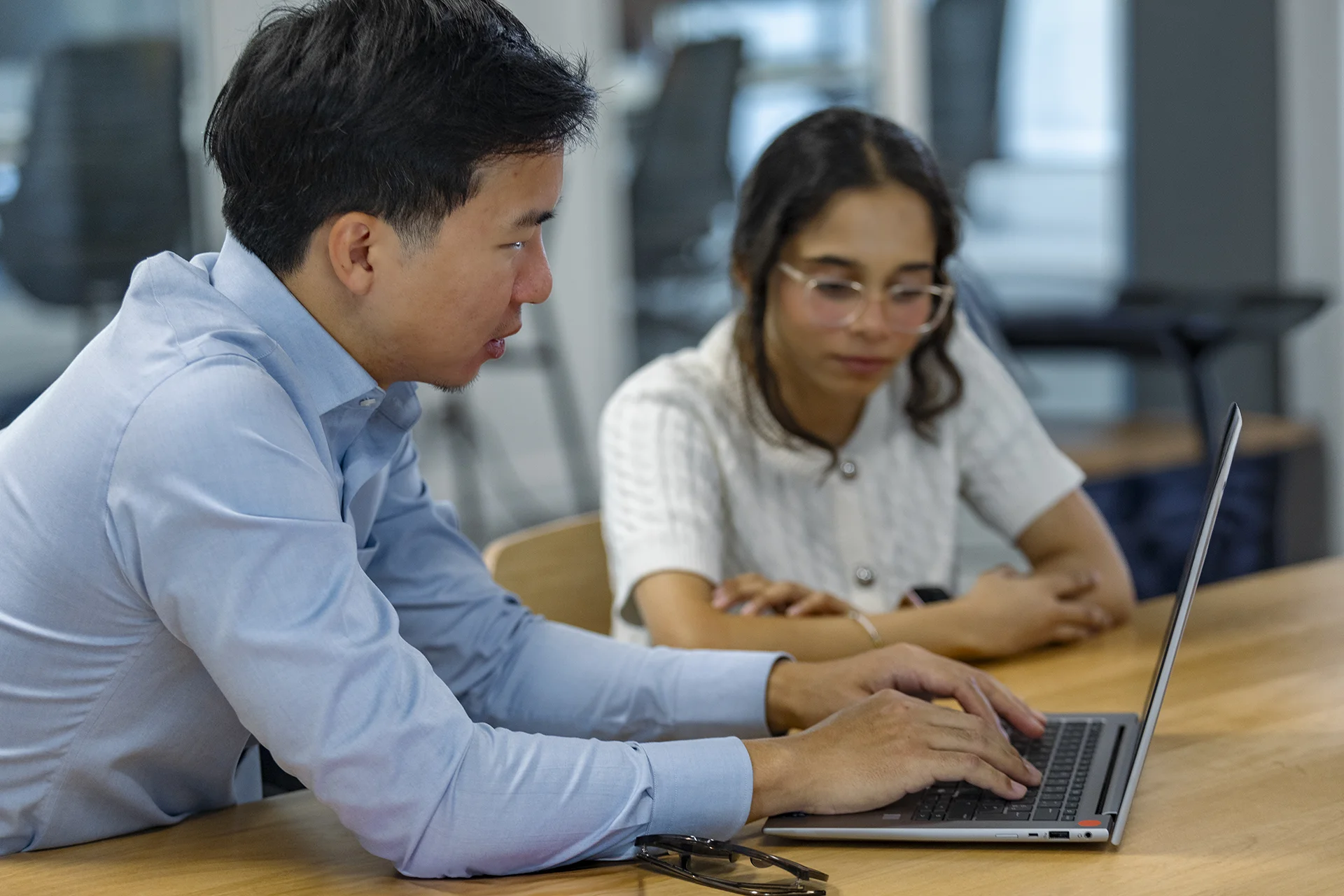 wo people seated at a wooden table working together on a laptop in a modern office setting. One person is typing on the keyboard while the other observes, creating a collaborative work atmosphere