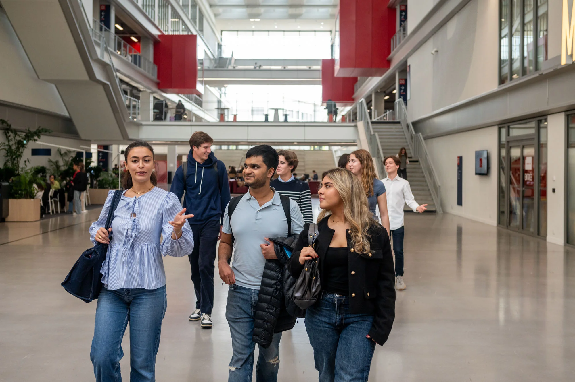 Un groupe d’étudiants et d’étudiantes d’emlyon marche dans le vaste atrium lumineux de l’école. L’espace présente de grandes baies vitrées, des passerelles, des escaliers et des éléments architecturaux rouges qui structurent le bâtiment. Le groupe avance au premier plan en discutant, tandis que d’autres personnes se déplacent à l’arrière-plan dans le cœur battant du campus.