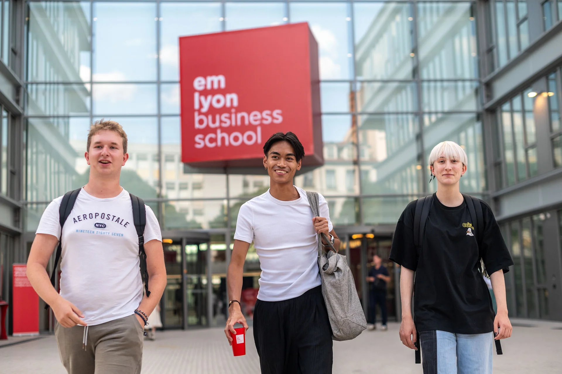 Étudiants marchant devant l’entrée d'emlyon business school sur le campus, devant le bâtiment principal au logo rouge.