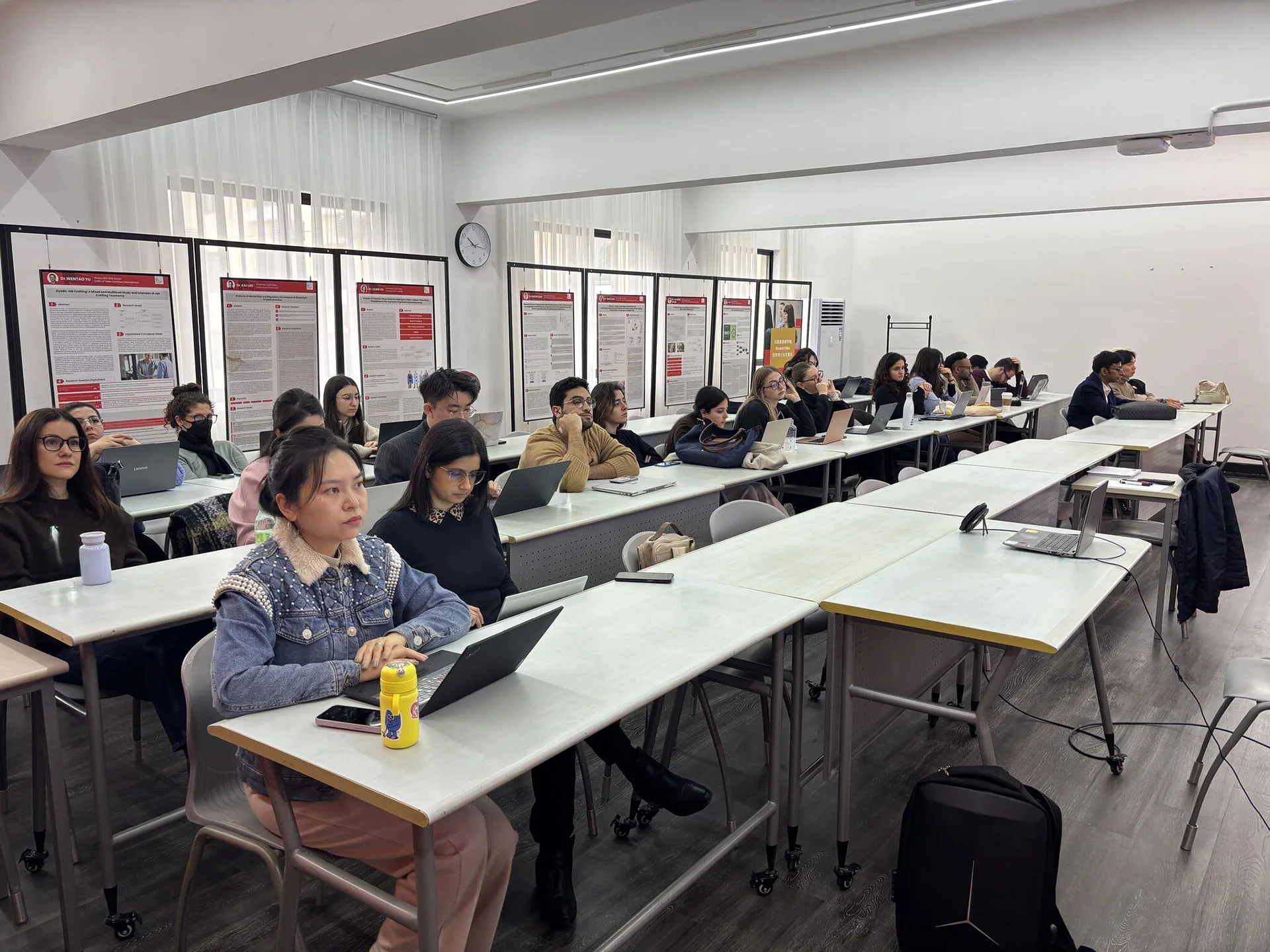 Un groupe d'étudiants attentifs dans un classroom moderne, assis à des tables en rangée avec des ordinateurs portables ouverts devant eux. Des affiches informatives se trouvent en arrière-plan, ajoutant un élément d'apprentissage visuel. L'atmosphère est studieuse et concentrée, mettant en avant l'engagement des étudiants dans leur éducation.