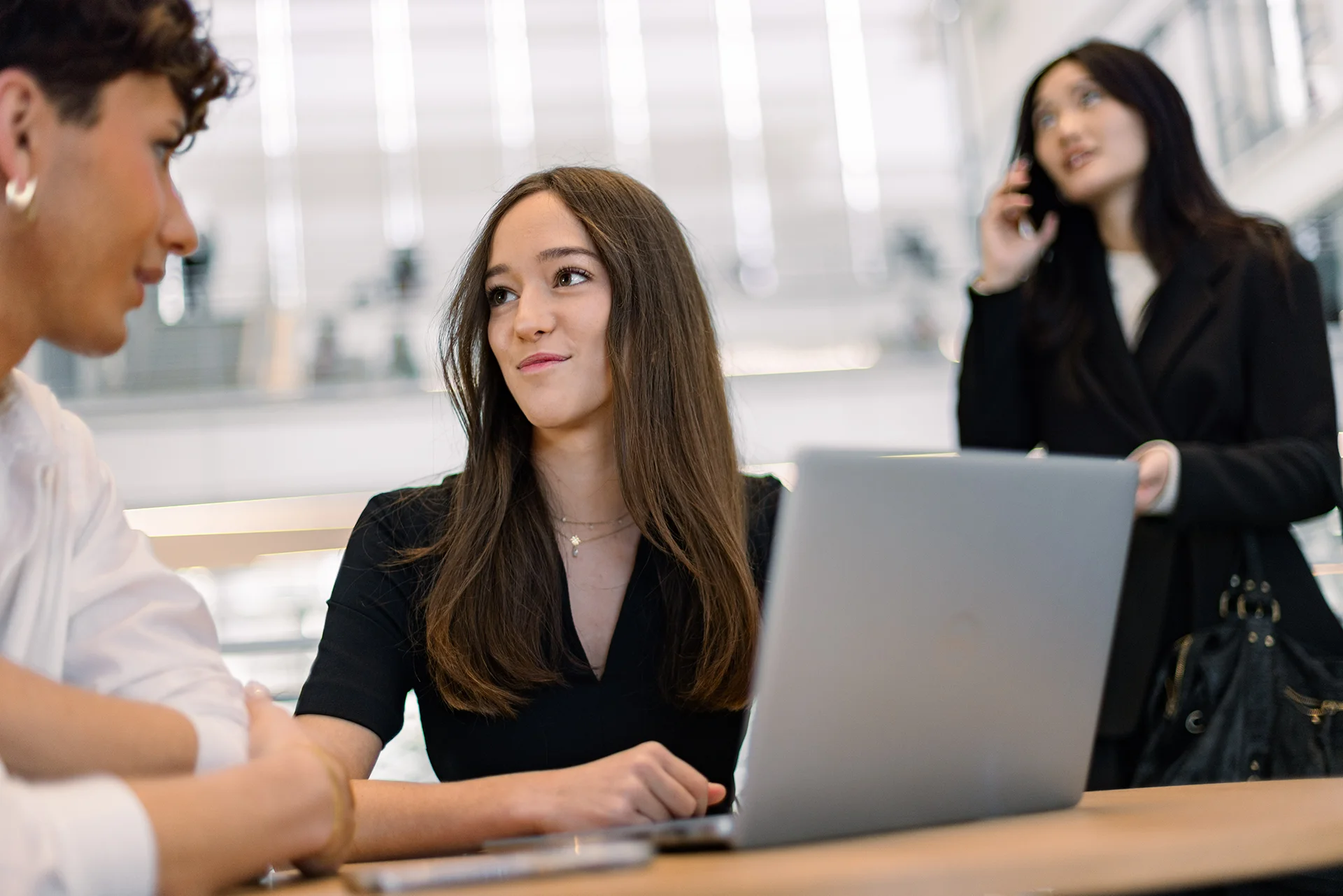 Trois personnes dans un espace de travail moderne. Une femme au premier plan porte un haut noir et travaille sur un ordinateur portable, tandis qu'une autre femme en arrière-plan est au téléphone, et un homme est à côté d'elle, portant une chemise blanche. L'environnement est lumineux et contemporain, avec des éléments architecturaux visibles.
