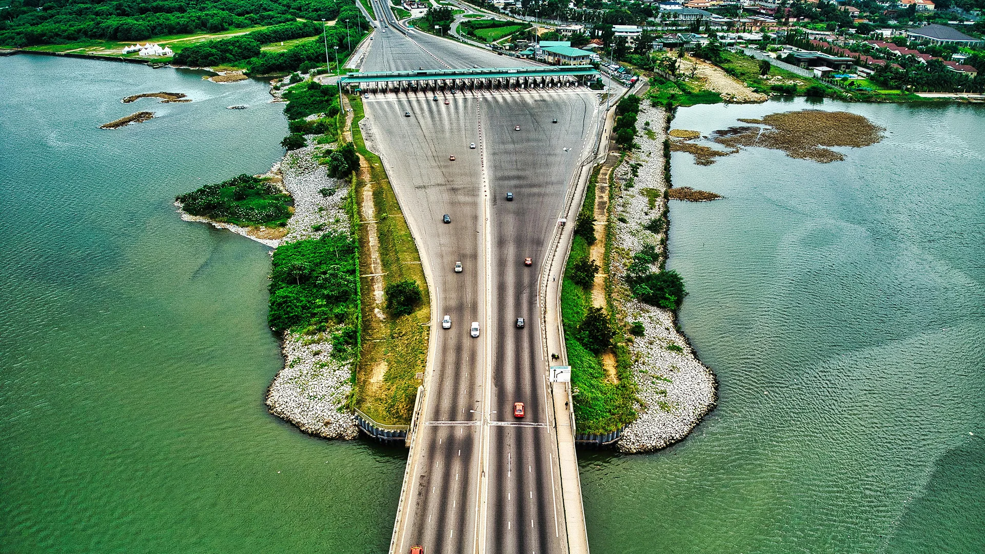 Vue aérienne d’un pont routier à plusieurs voies traversant une lagune à Abidjan, avec un péage, des véhicules en circulation et des berges végétalisées.