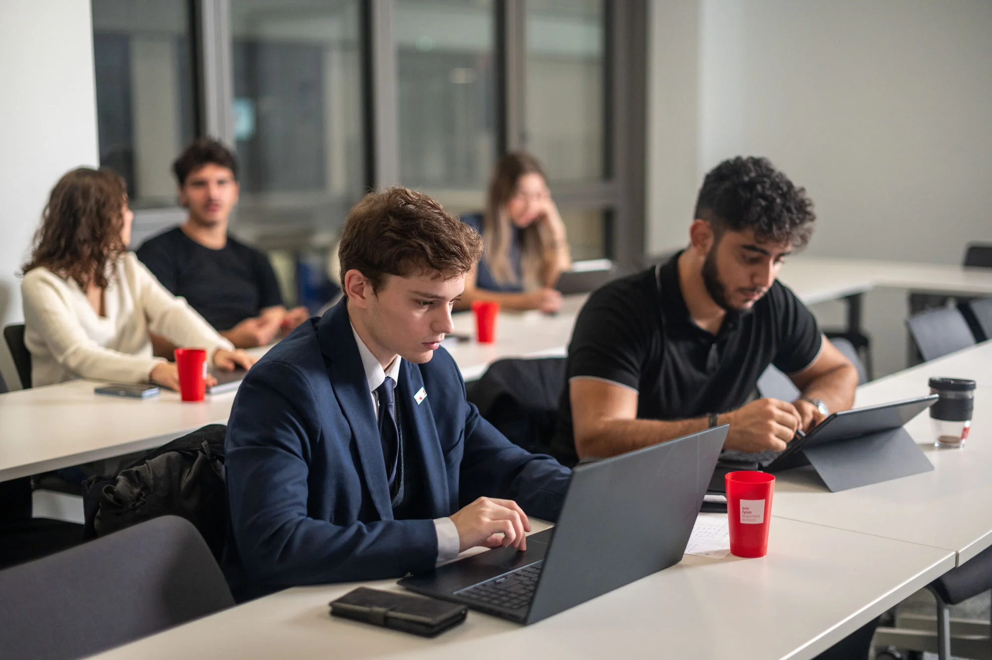 IMBA students at emlyon business school working on laptops during a class in a modern glass-walled classroom.