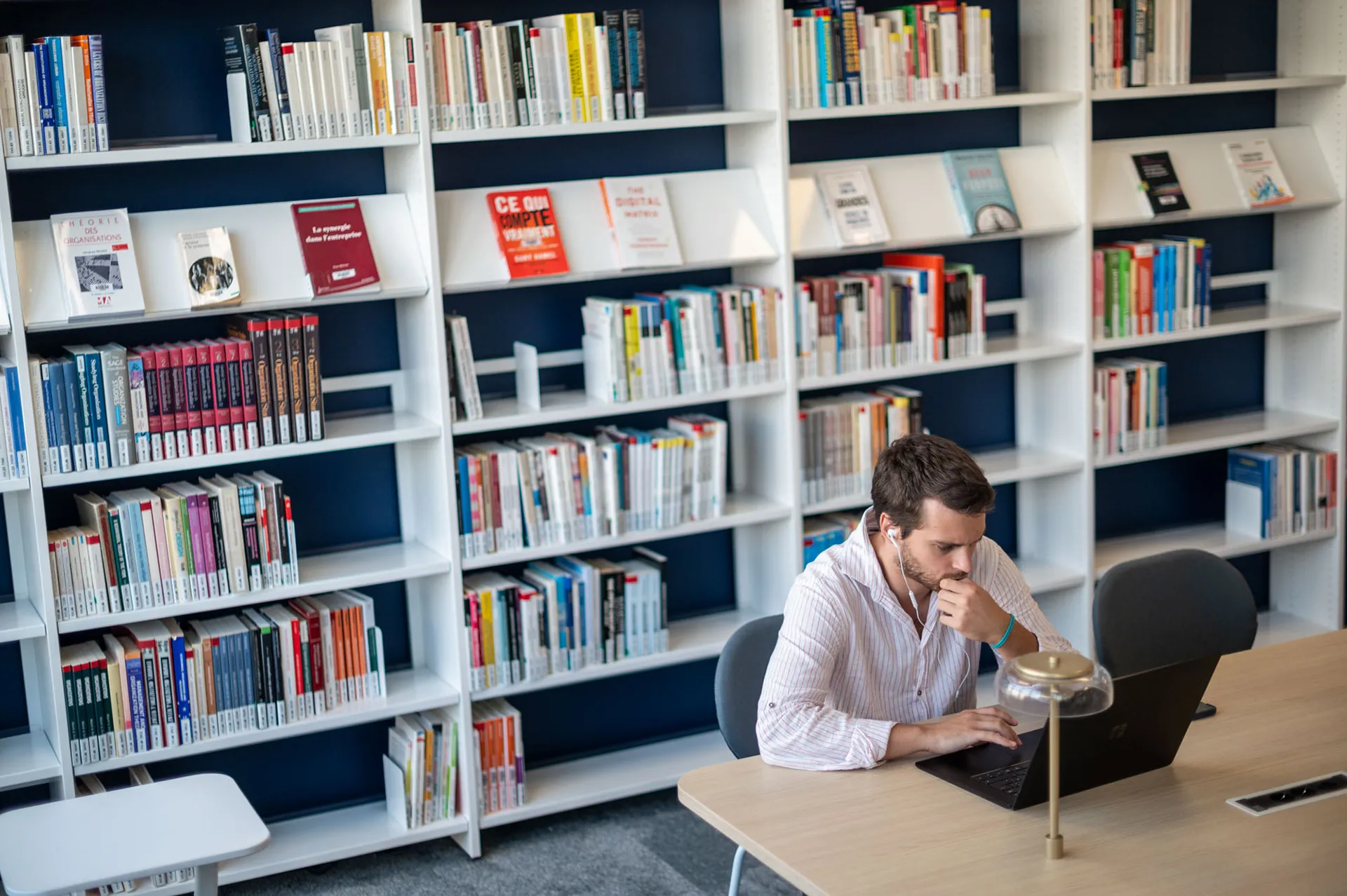 A person is seated at a table in a library, using a laptop computer, with bookshelves in the background and a desk lamp on the table.