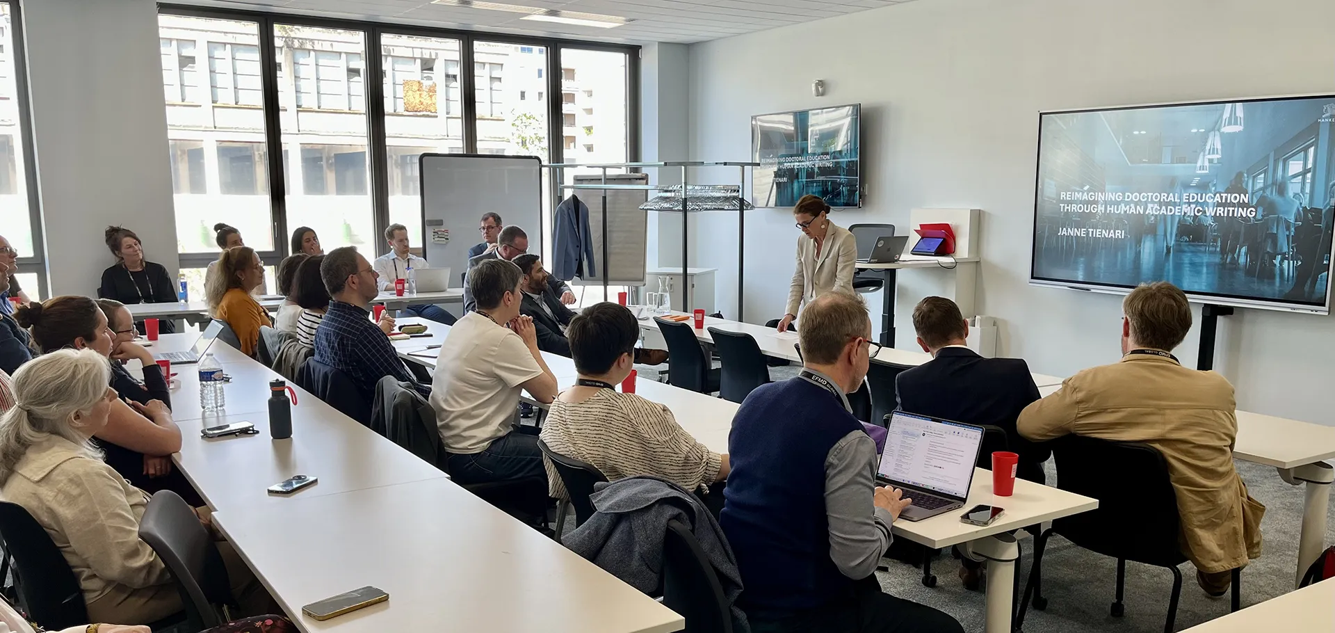 Conference room with people seated at tables while a presentation is shown on a screen.