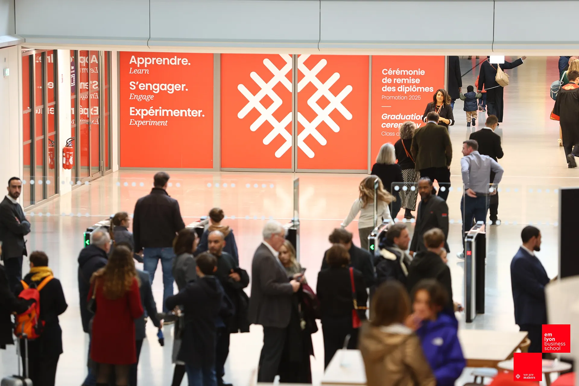 People entering and walking through a university campus hall; orange panels display the words “Learn”, “Engage”, “Experiment” and information about a graduation ceremony.