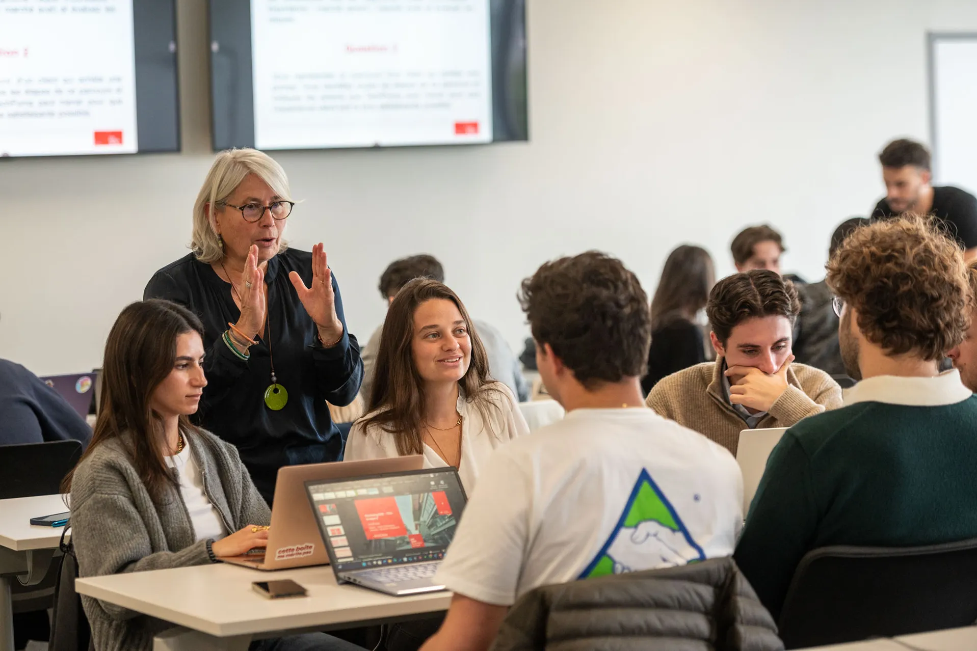 Classroom scene showing an instructor speaking with students gathered around laptops during a collaborative learning activity.