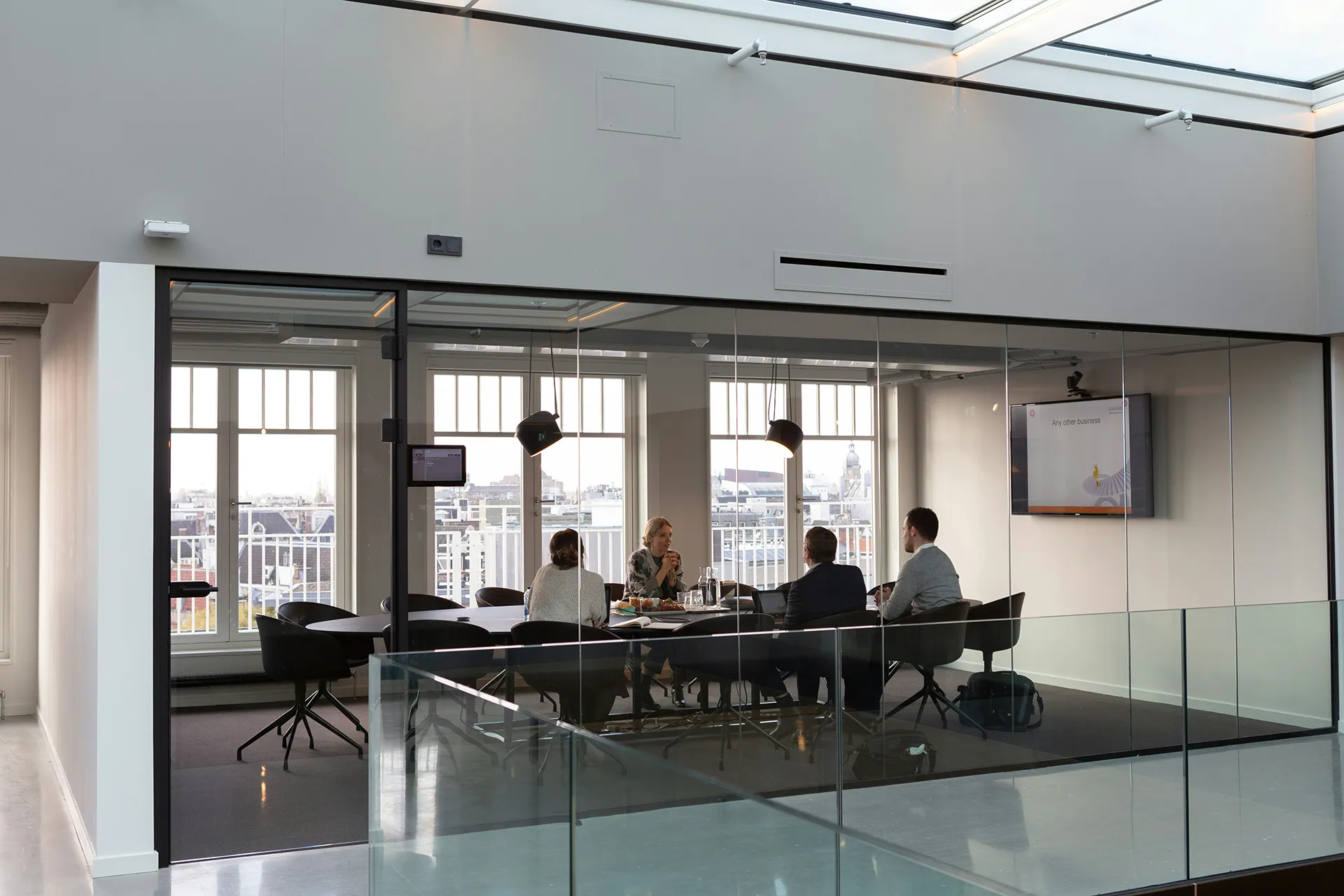 Salle de réunion vitrée avec une table centrale et plusieurs personnes assises, dans un espace de travail professionnel lumineux.