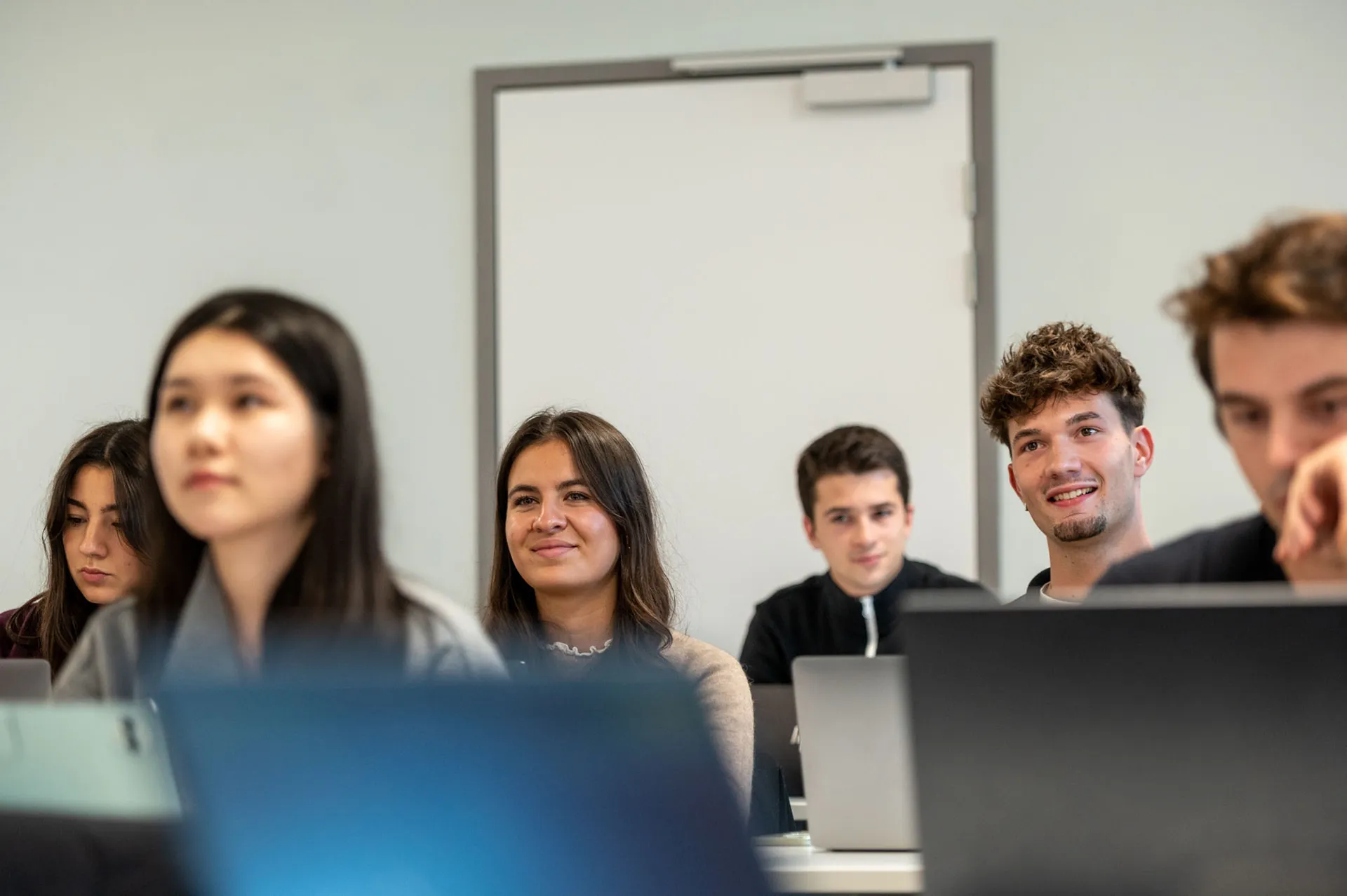 Un groupe de personnes assises dans une salle de classe utilise des ordinateurs portables, avec une porte fermée visible au fond de la pièce.