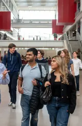 Un groupe d’étudiants et d’étudiantes d’emlyon marche dans le vaste atrium lumineux de l’école. L’espace présente de grandes baies vitrées, des passerelles, des escaliers et des éléments architecturaux rouges qui structurent le bâtiment. Le groupe avance au premier plan en discutant, tandis que d’autres personnes se déplacent à l’arrière-plan dans le cœur battant du campus.