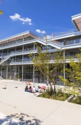 Vue du campus emlyon business school à Lyon, montrant un bâtiment moderne à plusieurs niveaux avec terrasses et passerelles extérieures, entouré d’arbres et d’espaces paysagers. Au premier plan, quelques personnes sont assises sur une esplanade en plein soleil.