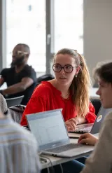 Student in a classroom working on a laptop during an in‑person lesson, in a bright room with large windows.