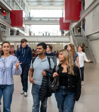 Un groupe d’étudiants et d’étudiantes d’emlyon marche dans le vaste atrium lumineux de l’école. L’espace présente de grandes baies vitrées, des passerelles, des escaliers et des éléments architecturaux rouges qui structurent le bâtiment. Le groupe avance au premier plan en discutant, tandis que d’autres personnes se déplacent à l’arrière-plan dans le cœur battant du campus.