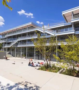 Vue du campus emlyon business school à Lyon, montrant un bâtiment moderne à plusieurs niveaux avec terrasses et passerelles extérieures, entouré d’arbres et d’espaces paysagers. Au premier plan, quelques personnes sont assises sur une esplanade en plein soleil.