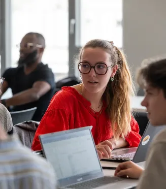 Student in a classroom working on a laptop during an in‑person lesson, in a bright room with large windows.