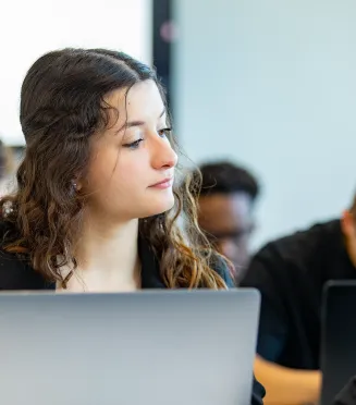 Étudiant·e assis·e dans une salle de classe, utilisant un ordinateur portable, avec d’autres étudiants travaillant en arrière-plan.