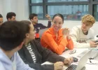 Students collaborating around laptops in a classroom, with one person in an orange shirt at the center engaging with peers