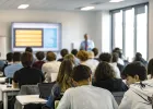 People are seated at tables in a lecture hall, some using laptops, while a presentation is projected onto a screen at the front of the room.