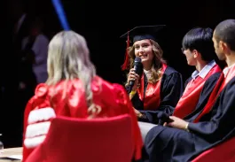 Cérémonie de remise de diplômes : une diplômée en toge noire et écharpe rouge parle au micro sur scène, aux côtés d’autres diplômés et d’un intervenant.