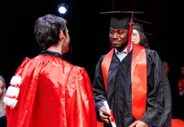 Remise de diplômes : un diplômé en toge noire et écharpe rouge reçoit son diplôme des mains d’un représentant sur scène.