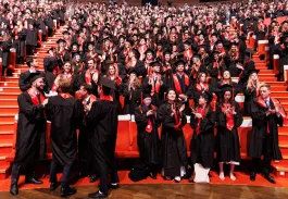 Grand amphithéâtre rempli de diplômés en toge noire et écharpe rouge applaudissant lors d’une cérémonie de remise de diplômes.
