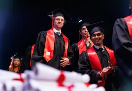 Diplômés en toge noire et écharpe rouge debout sur scène lors d’une cérémonie de remise de diplômes, avec des diplômes au premier plan.