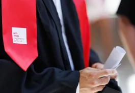 Gros plan d’un diplômé en toge noire avec écharpe rouge tenant un diplôme, logo emlyon business school visible.