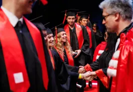 Remise de diplômes : des diplômés en toge noire et écharpe rouge serrent la main d’un représentant sur scène.