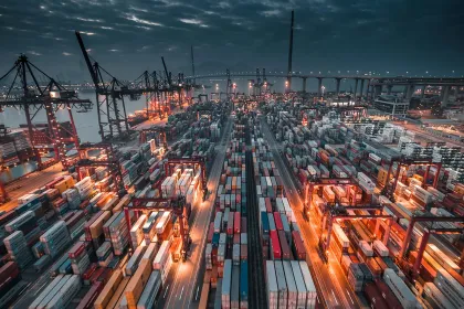 Vue aérienne d'un port de conteneurs de nuit, avec de nombreuses cargaisons colorées empilées, des grues surélevées et des lumières brillantes qui illuminent la zone de chargement.