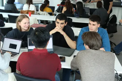 L'image montre un groupe de personnes rassemblées autour d'une table, travaillant sur des ordinateurs portables. Ils semblent être engagés dans une discussion ou une session de collaboration. Au fond, d'autres personnes et ordinateurs sont visibles, suggérant un environnement de travail ou d'apprentissage collaboratif. La scène dégage une atmosphère de convivialité et d'interaction.