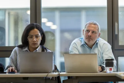 Deux personnes assises à une table de réunion, travaillant sur leurs ordinateurs portables. Un café et un verre d'eau sont présents sur la table, avec une grande fenêtre en arrière-plan laissant passer la lumière.