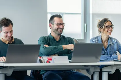 Groupe de trois personnes assises à une table, avec des ordinateurs portables, dans un cadre professionnel moderne. Ils semblent engagés dans une discussion ou une réunion.