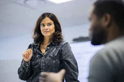 Une femme est debout, en train de parler, vêtue d'une chemise en jean. Elle semble engager une conversation ou présenter quelque chose, avec une main levée. En arrière-plan, on aperçoit un tableau ou un écran, suggérant un cadre de réunion ou de présentation. La scène dégage une atmosphère d'interaction professionnelle ou éducative.