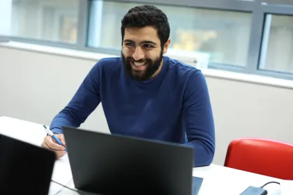 Un homme assis à une table, portant un pull bleu. Il tient un stylo et semble écrire. Sur la table, il y a un ordinateur portable noir et un autre appareil. Une fenêtre se trouve en arrière-plan.