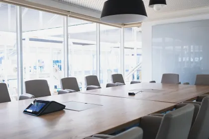 Salle de réunion lumineuse avec une grande table en bois, des chaises modernes disposées autour, un dispositif de visioconférence et de larges baies vitrées laissant entrer la lumière naturelle.
