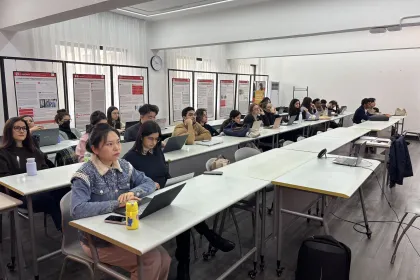 Un groupe d'étudiants attentifs dans un classroom moderne, assis à des tables en rangée avec des ordinateurs portables ouverts devant eux. Des affiches informatives se trouvent en arrière-plan, ajoutant un élément d'apprentissage visuel. L'atmosphère est studieuse et concentrée, mettant en avant l'engagement des étudiants dans leur éducation.