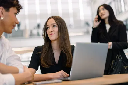Trois personnes dans un espace de travail moderne. Une femme au premier plan porte un haut noir et travaille sur un ordinateur portable, tandis qu'une autre femme en arrière-plan est au téléphone, et un homme est à côté d'elle, portant une chemise blanche. L'environnement est lumineux et contemporain, avec des éléments architecturaux visibles.