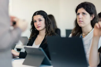 Personnes assises dans une salle de cours moderne, utilisant des ordinateurs portables et une tablette, attentives à une intervention en présentiel, dans un contexte d’enseignement supérieur et de formation.