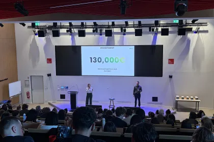 Vue de l'amphithéâtre d'emlyon rempli de participants assistant à une présentation. Deux intervenants se tiennent sur scène devant un grand écran indiquant « Investment – 130 000 € », avec un pupitre et un tabouret visibles.